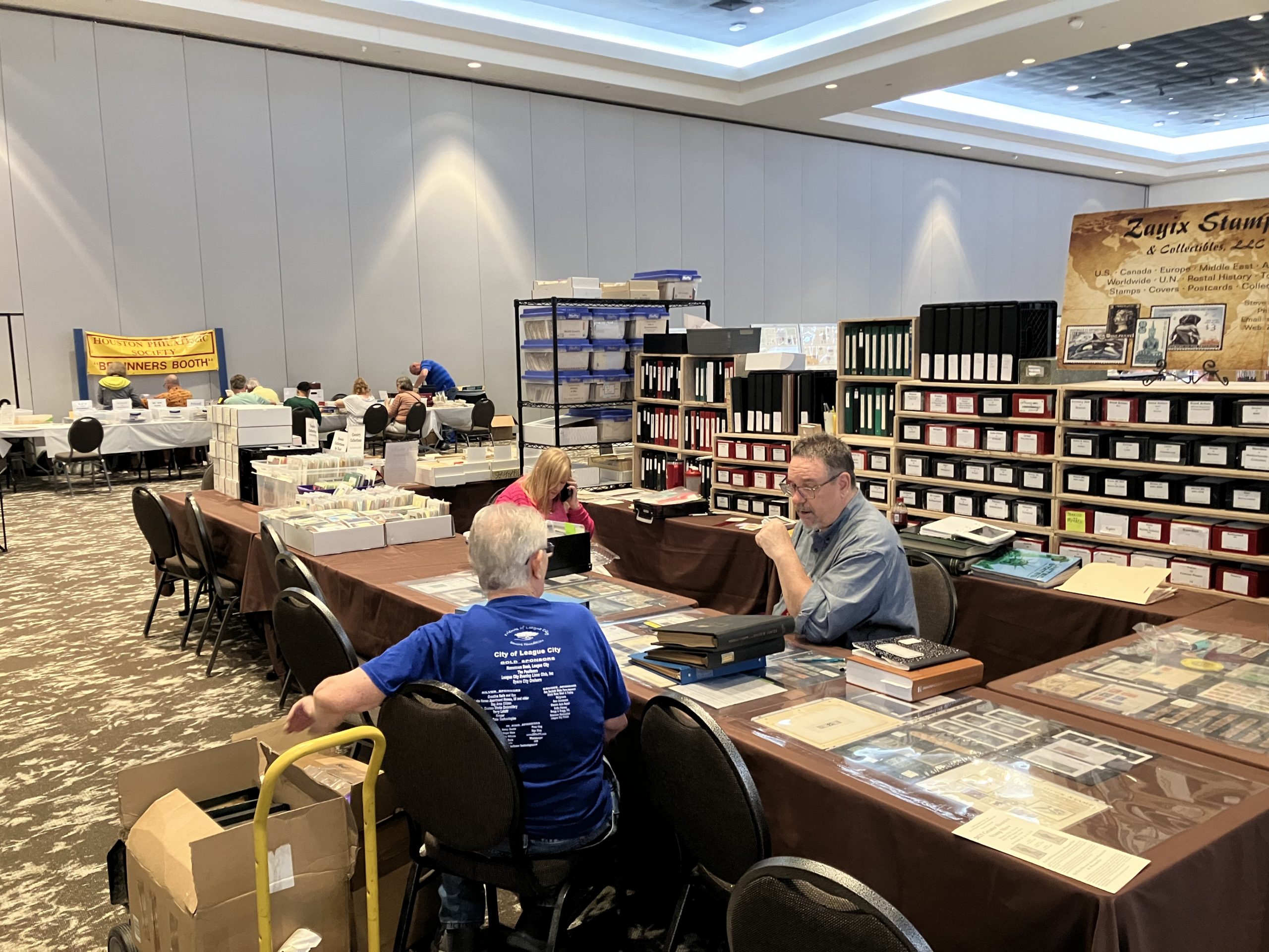 Steve Stewart from Zayix Stamps & Collectibles, LLC, talks stamps with a customer while his wife, Theresa, assists a customer on the phone. The Beginner's Booth can be seen to the left.